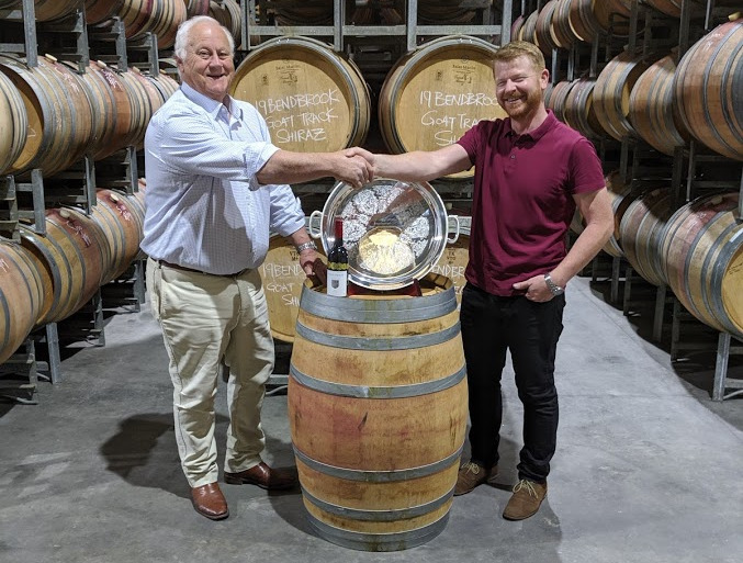 Two men shake hands over a trophy plate and a bottle of wine on top of an oak cask. Surrounding them are more oak casks on stands. The casks behind them have been marked "19 Bendbrook Goat Track Shiraz".