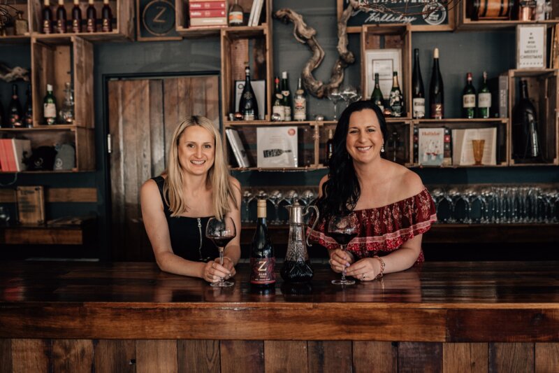 Two women behind a wooden bar, each holding a glass of dark red wine. Between them on the bar is a bottle of Z Julius Shiraz and a fancy carafe of wine with a metal top and handle. Behind them is bar shelving holding glasses, bottles, and books.