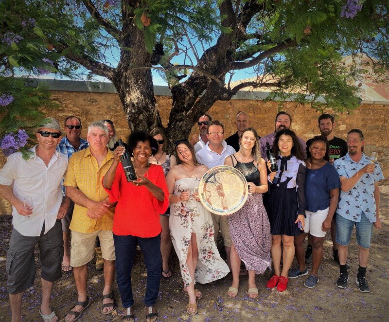 A group of 16 happy people shelter under the shade of a jacaranda tree in front of a sandstone block building. Two women in the centre of the group are holding a trophy plate, and women on either side of them each hold a bottle of Kirrihill wine. Jacaranda flowers litter the dry ground below.