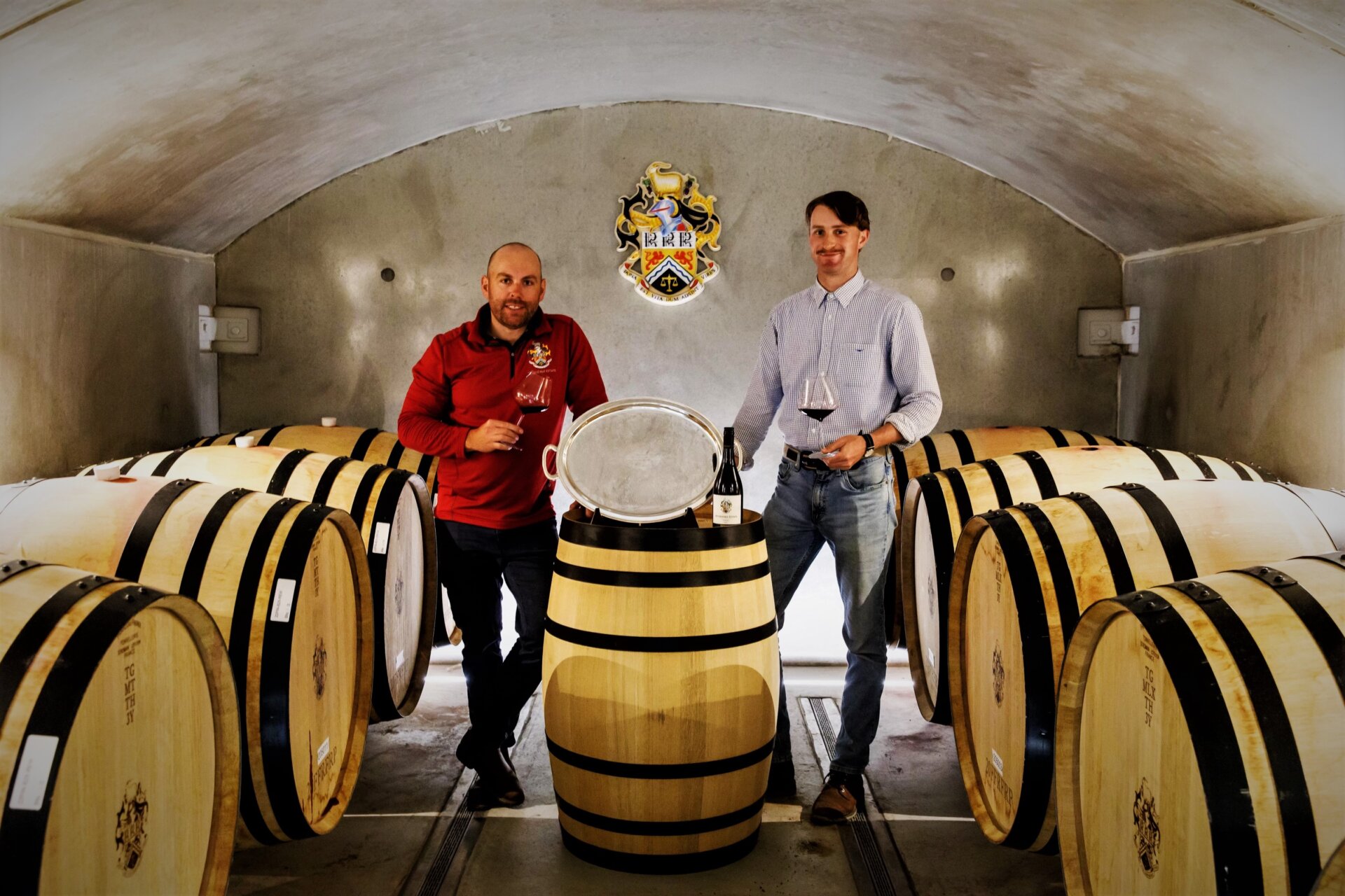 Two men stand either side of a wine barrel in a room with other wine barrels. There is a trophy plate displayed on the top of the wine barrel. Each man holds a glass of dark red wine.