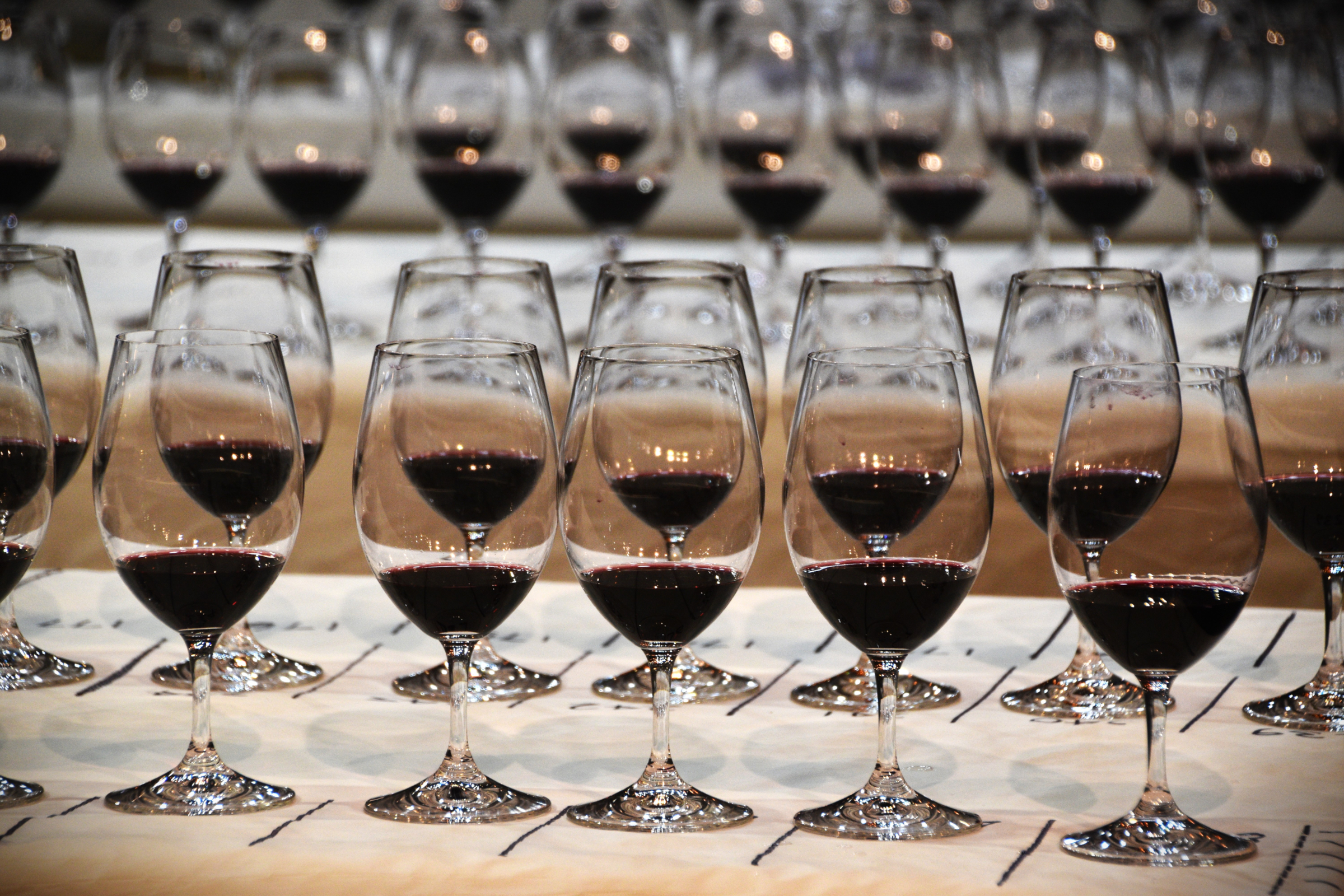 Rows of wine glasses with a small amount of red wine in each, sitting on thin tables between black lines marked on white paper. Each glass has a number on the table. Tables blur into the distance.
