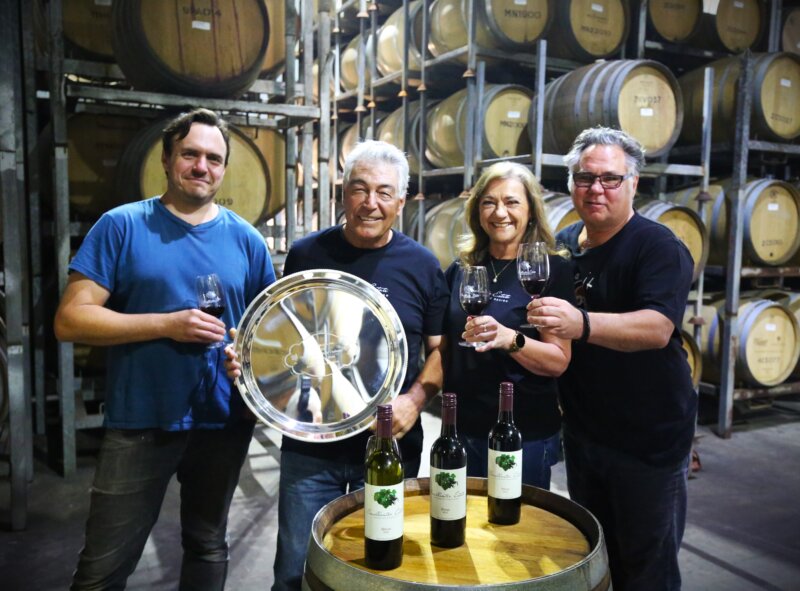 The Smallwater Team. Four people stand behind a wine barrel bearing three bottles of red wine. One holds a trophy plate while the others each hold a glass of red wine.